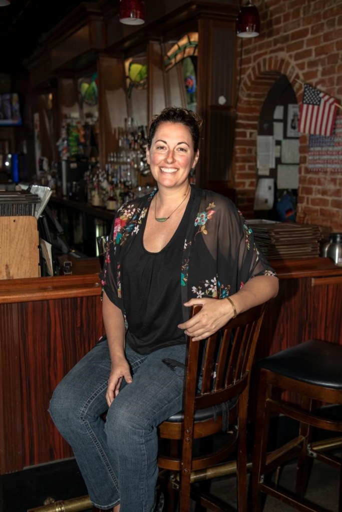 Photo of a smiling Christy, proudly sitting on a barstool in her restaurant. 