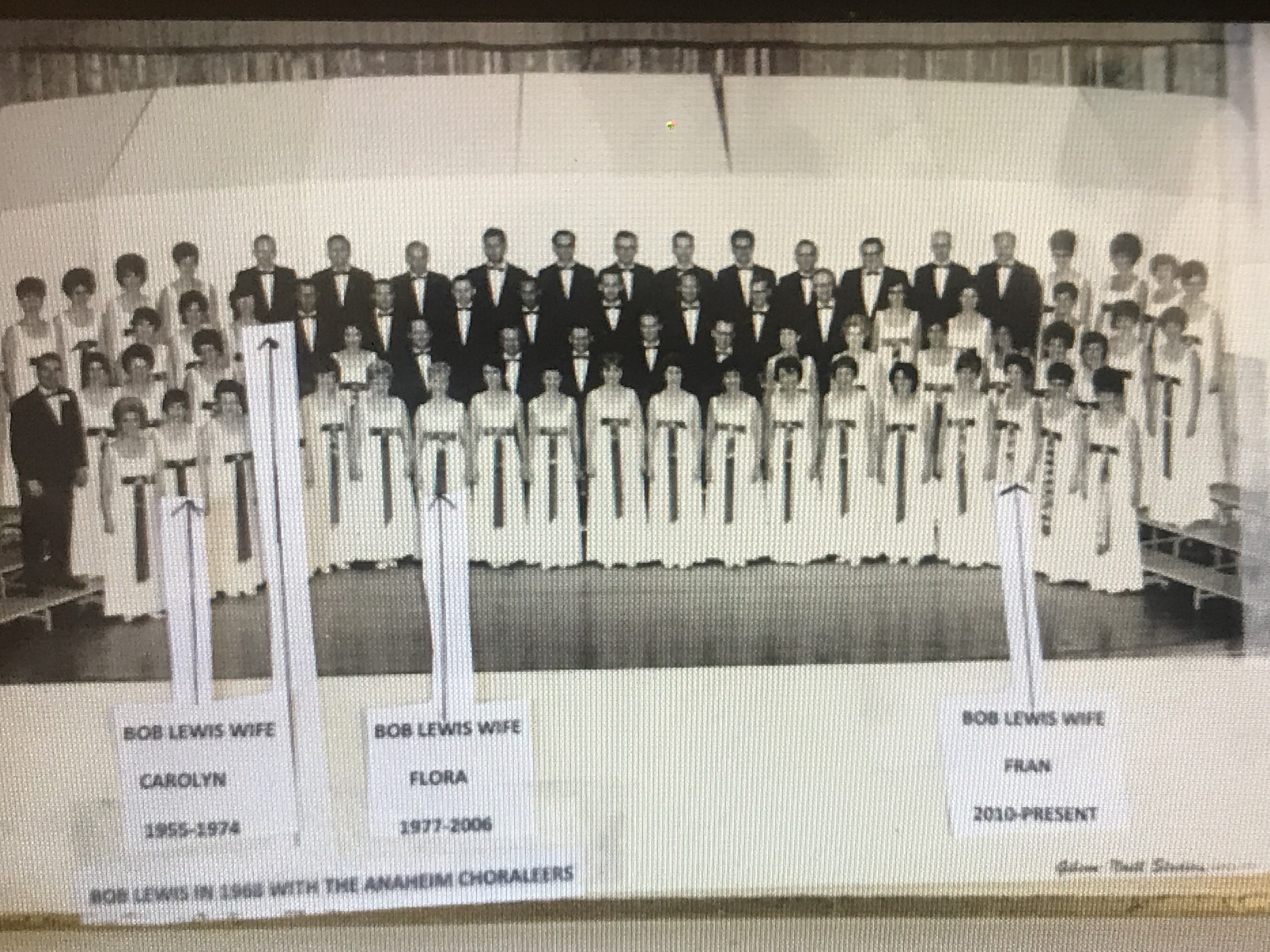 Formal portrait of 30-person Anaheim Choraleers. Men dressed in black tuxedos, women in floor length white gowns.