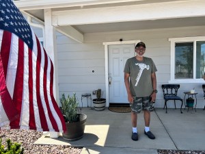 A man standing on front porch of house, with American flag at left.