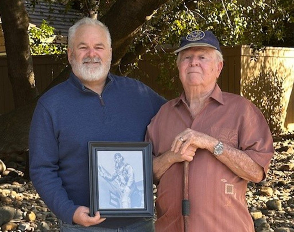 Two men standing outside, one holding a framed black and white photograph.